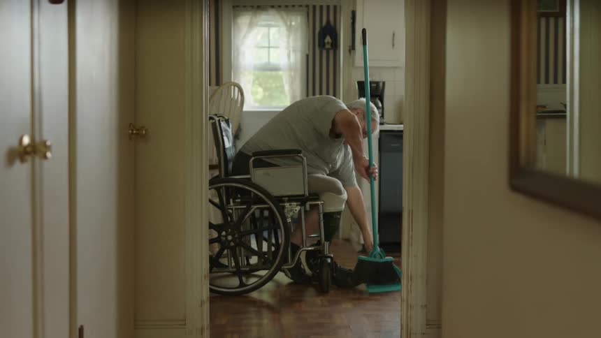 A man in a wheelchair sweeping the floor with a broom and shovel with a wall and window in the background