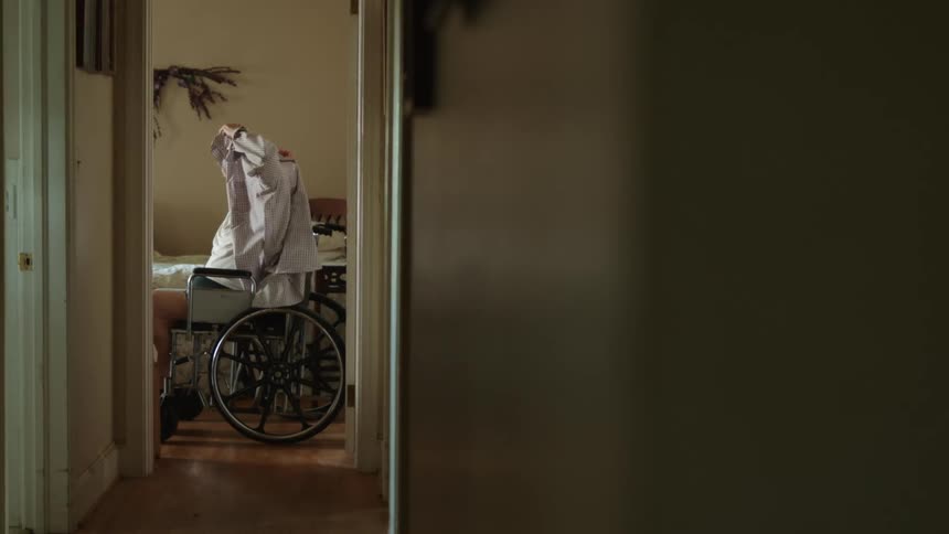 An elderly man sitting in a wheelchair puts his shirt on