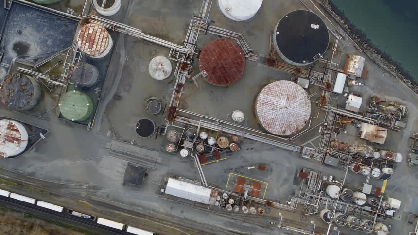 Top down view of tanks and pipework at a waterfront industrial facility as a train travels alongside