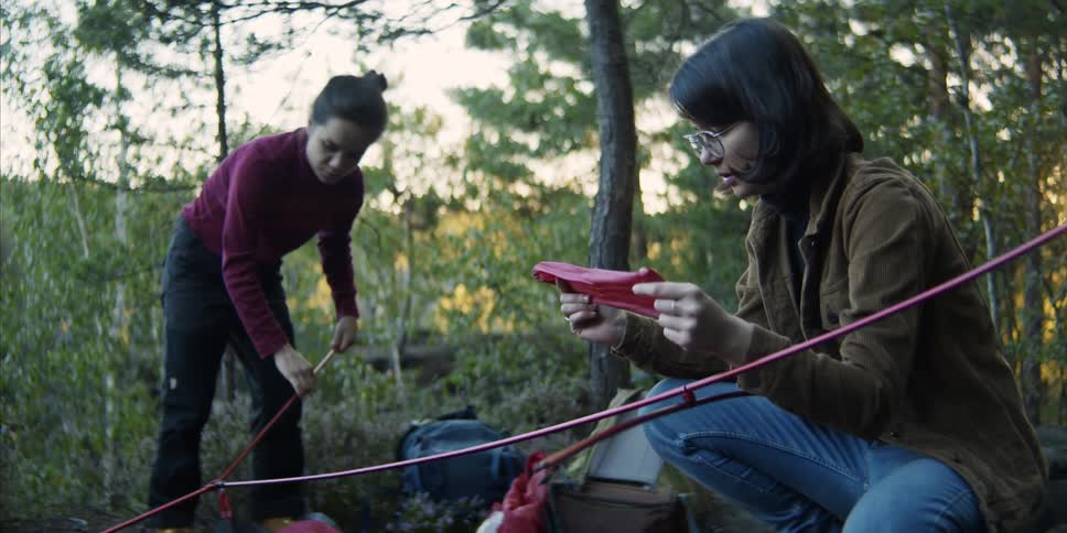 A teenage girl reads a paper while another teenage girl sets up a tent in a forest