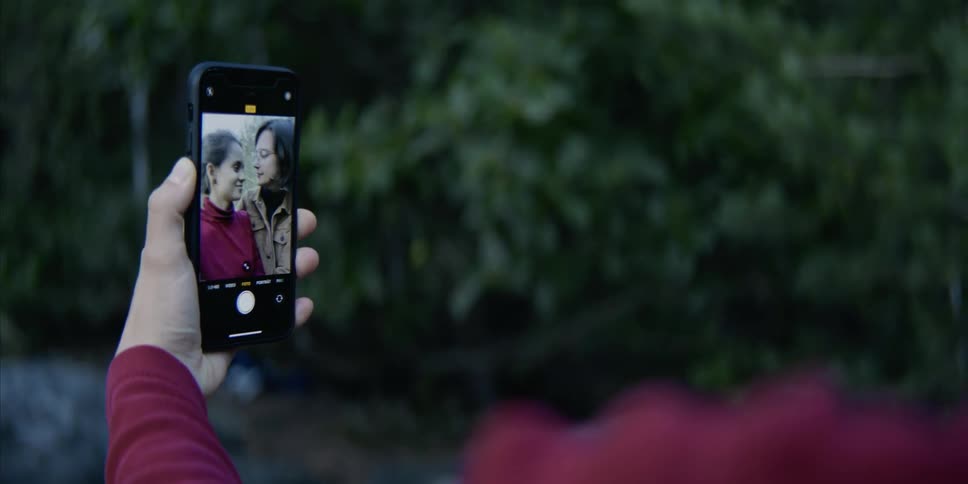 A woman uses a smartphone to record herself as she kisses another woman outdoors during the day