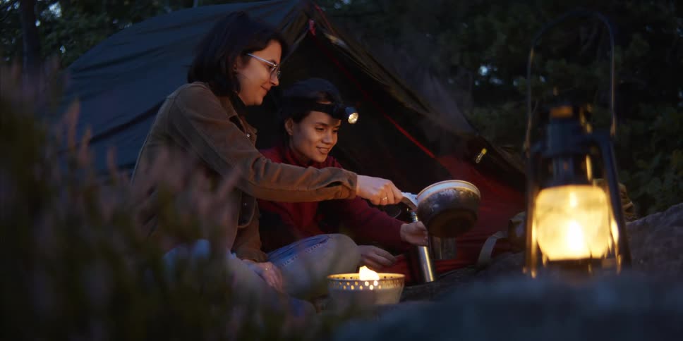A woman lifts a pot off a campfire and pours the contents into a cup being held by the woman sitting next to her as they sit together at a campsite in the evening