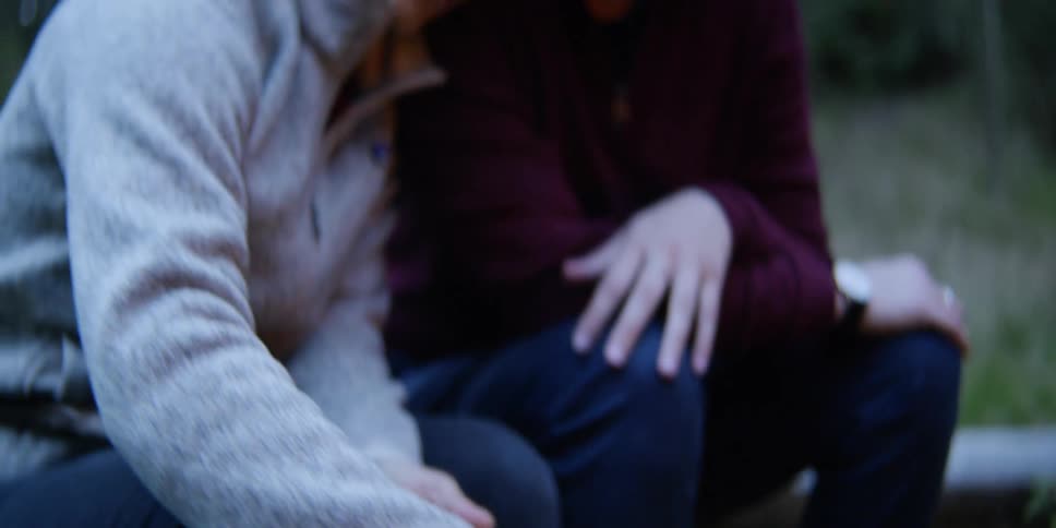 A young woman uses a knife to carve a stick into a sharp point as she rests her head against her partners shoulder as they sit on a log in a forest