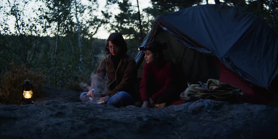 Two women sit on ground outside tent waiting for food to cook