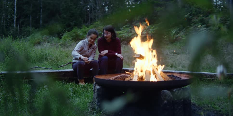 Two girls sit next to a fire while one whittles a stick