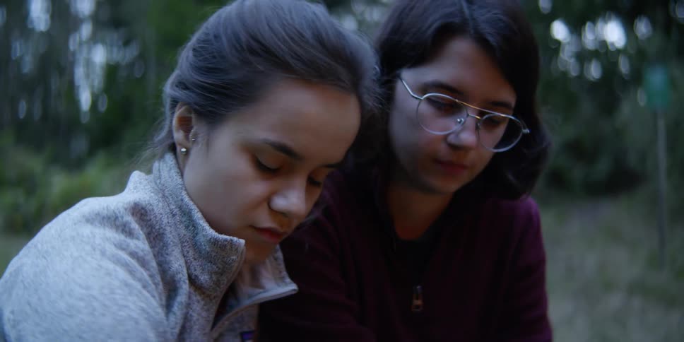 A pair of young women sit together outdoors in a front of a campfire