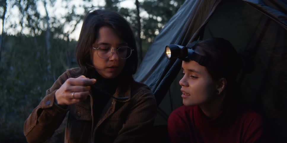A young woman sitting outdoors in front of a tent lifts a spoonful of food from a pot and blows on it before feeding it to the woman sitting beside her