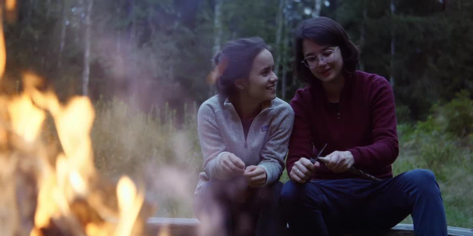Two women sit together on a log near a campfire as one whittles a stick