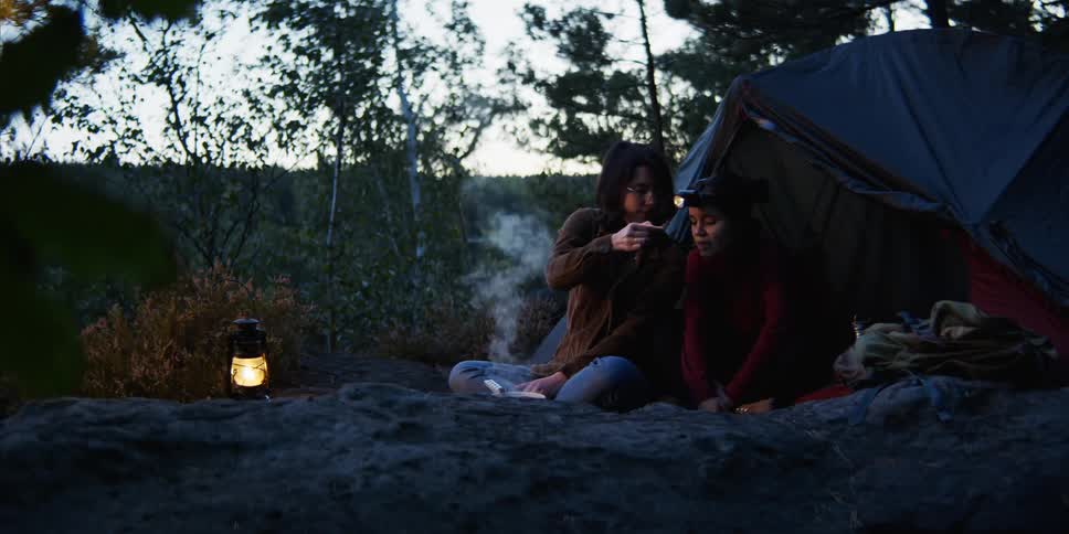 A woman cooking in front of tent feeds a spoonful of food to the woman sitting beside her before tasting it herself