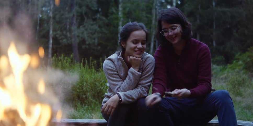 Two women sit around a campfire and one of them chips away at a stick with a knife