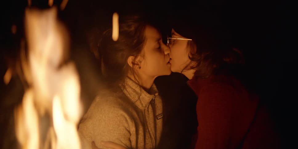 Two young women kiss while sitting outside by a campfire