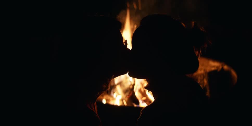 A pair of women illuminated only by firelight kiss outdoors at night