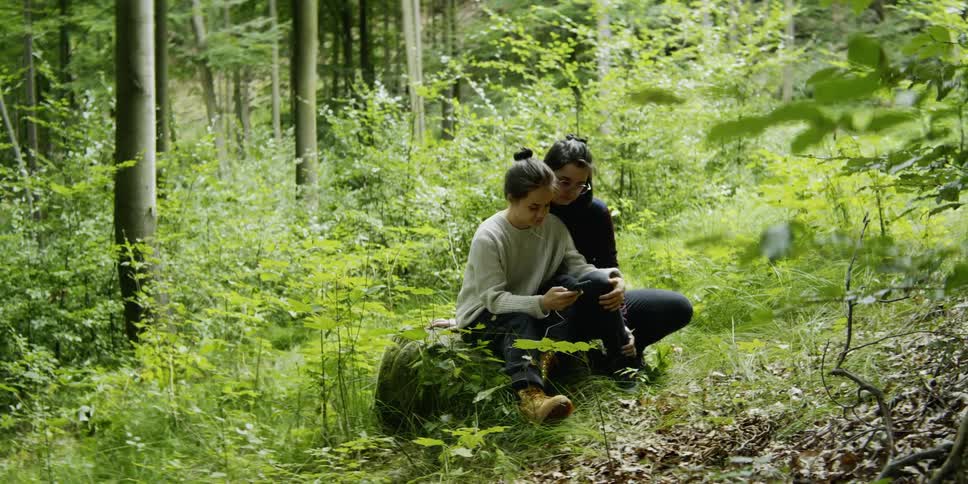 Two women sit in the woods looking at a phone