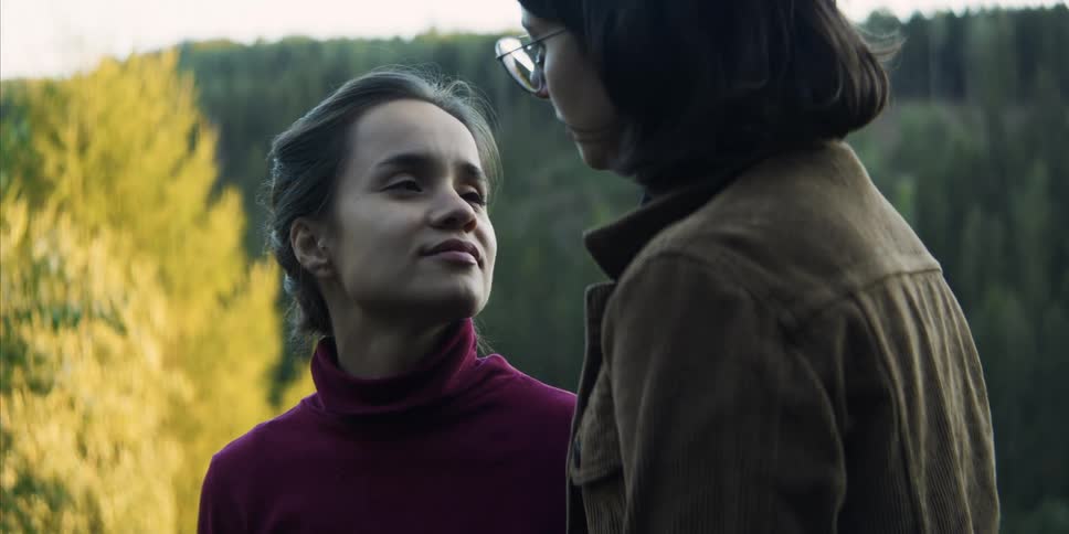 A young woman looking out at a forested countryside turns to share a kiss with the woman standing behind her