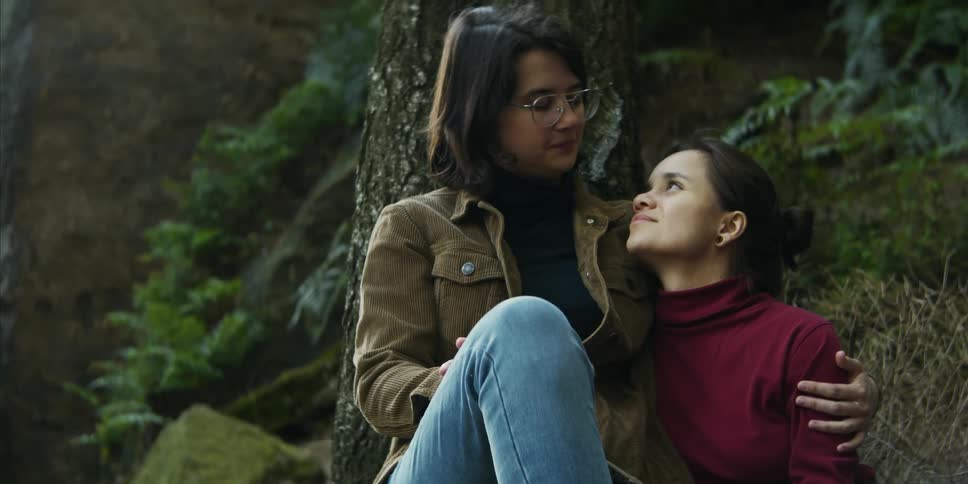 A young woman rubs her girlfriends shoulder as they kiss in a forest
