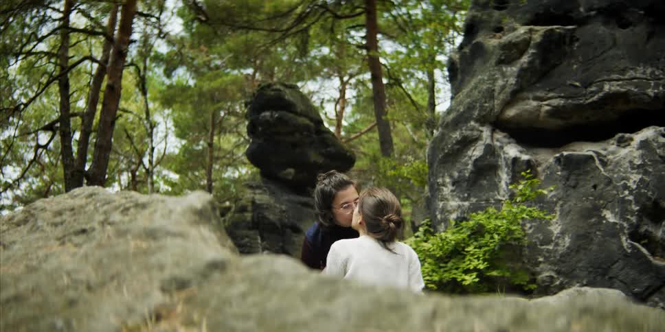 Lesbian couple with white and black sweaters kisses each other next to a big rocks in the woods
