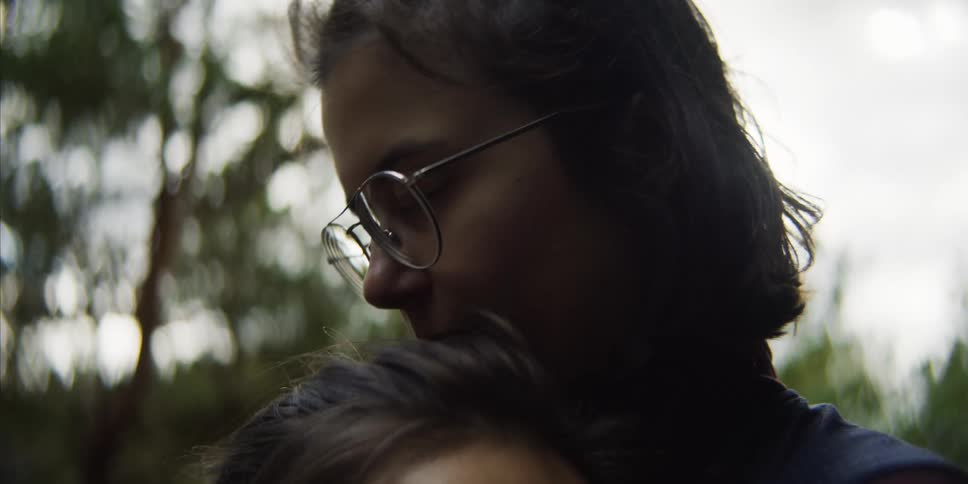 A woman kisses her girlfriend on the head as they are in a forest