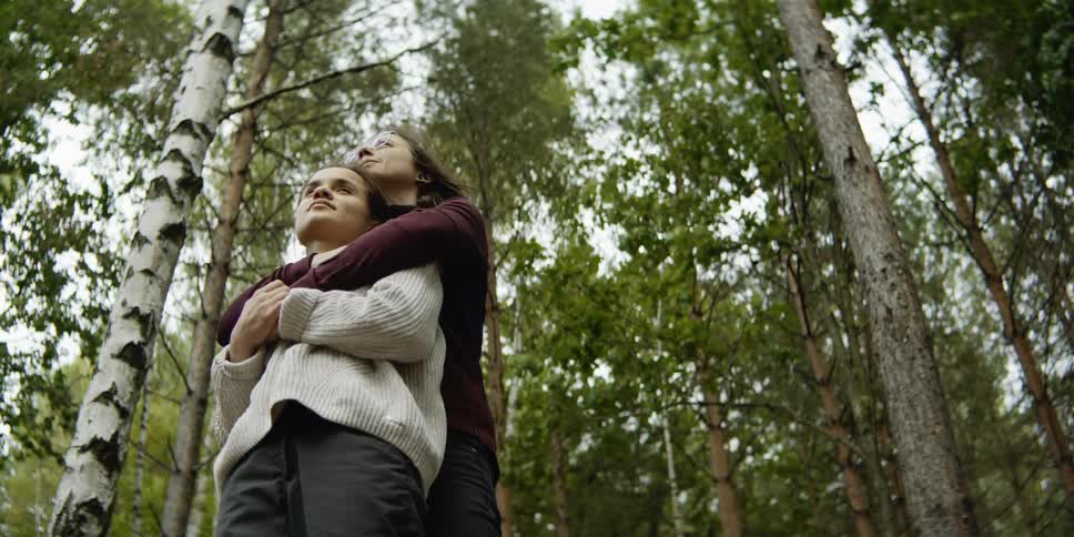 A young woman embraces from behind and kisses the head of another woman as they stand together in a forest during the day