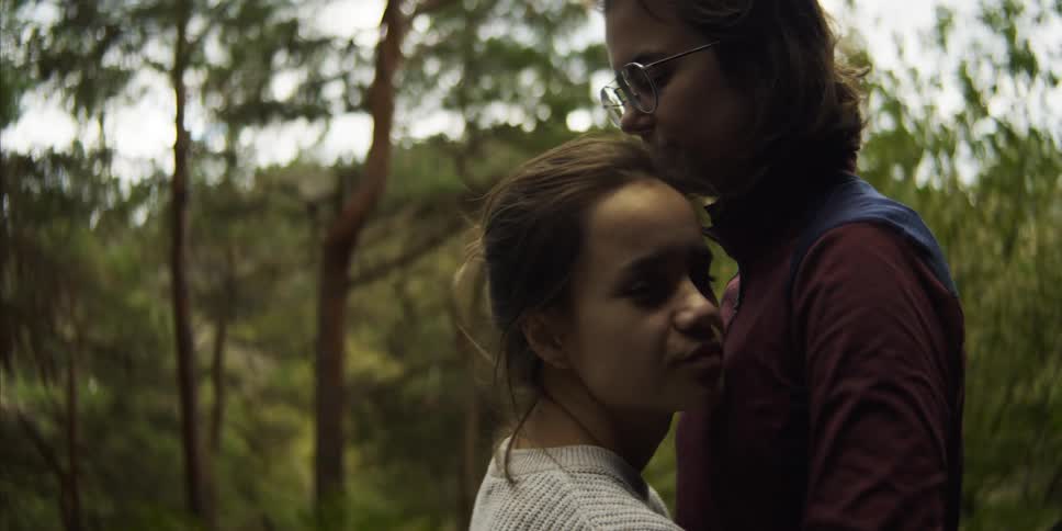 A pair of young women share a kiss in a woodland before embracing