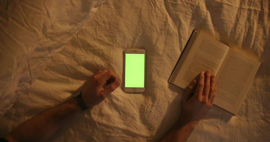 A man on a bed reads a book and scrolls on his phone