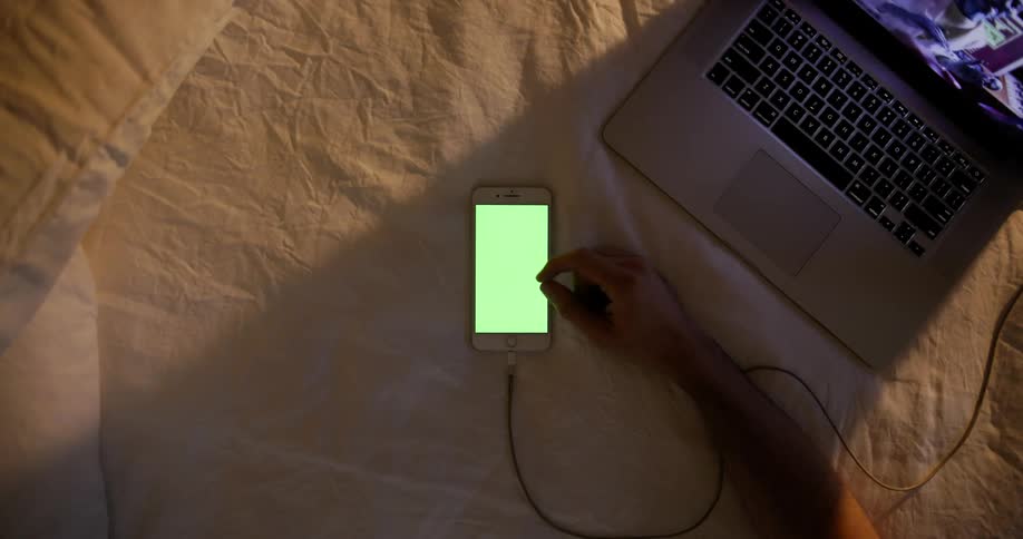 Man plays with smartphone on bed next to laptop