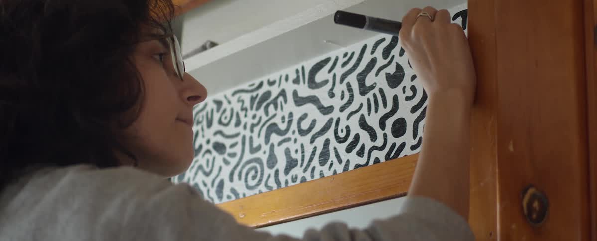 A woman with glasses and a marker in a room drawing a design on a wall