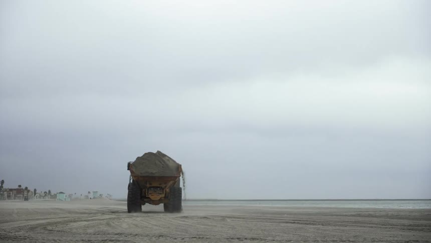 Yellow dump truck carries load of dirt off beach toward town under cloudy sky