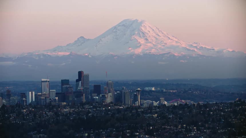 A stunning aerial of downtown Seattle at sunset with Mount Rainier in the background and skyscraper buildings in the foreground being illuminated by soft and golden sunset light