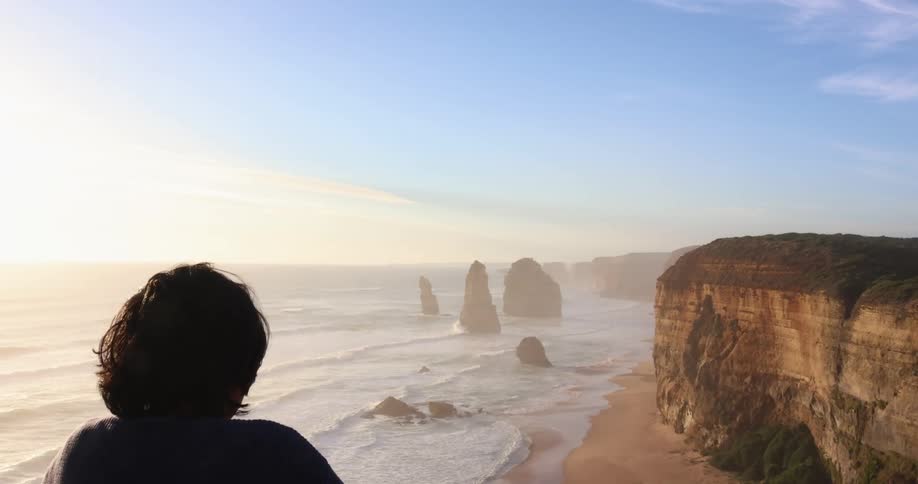 A boy sits on a cliff and look out at the waves rolling in to the shore