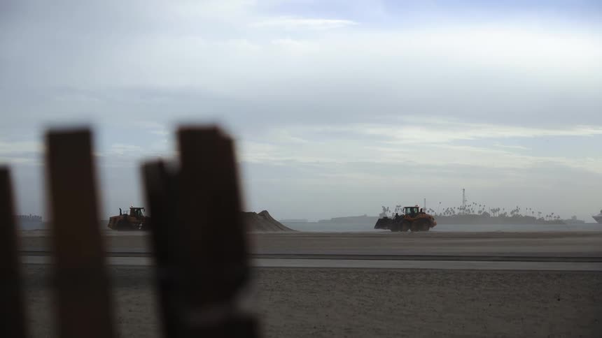 Two bulldozers parked on a windswept beach with a fence in the foreground