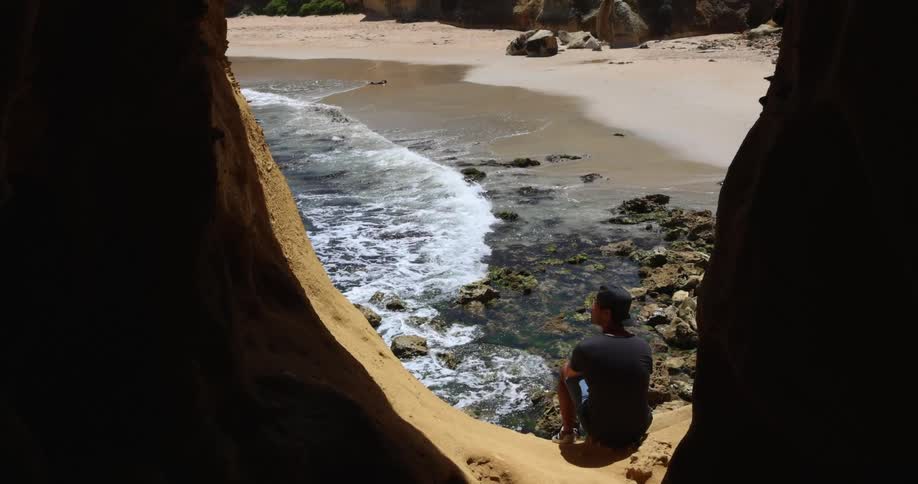 Man sits outside opening of cave near beach looking out to sea as waves crash