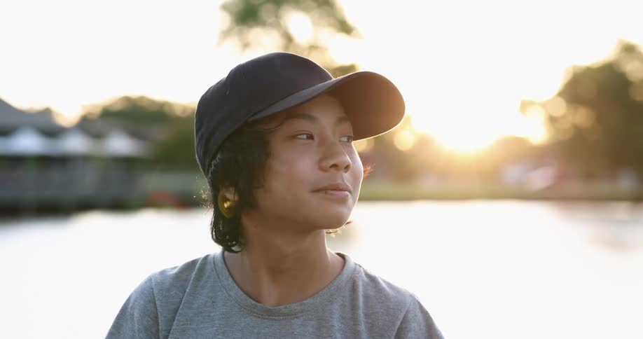 A teenager wearing a baseball cap stands in front of a lake at sunset