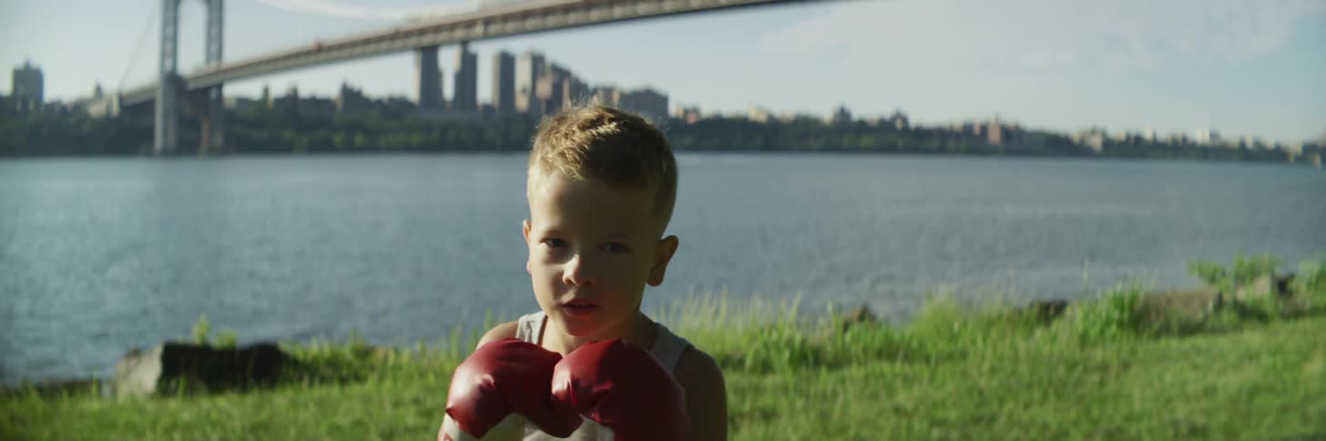 A young boy wearing boxing gloves shadow boxes on the river bank with a bridge behind him
