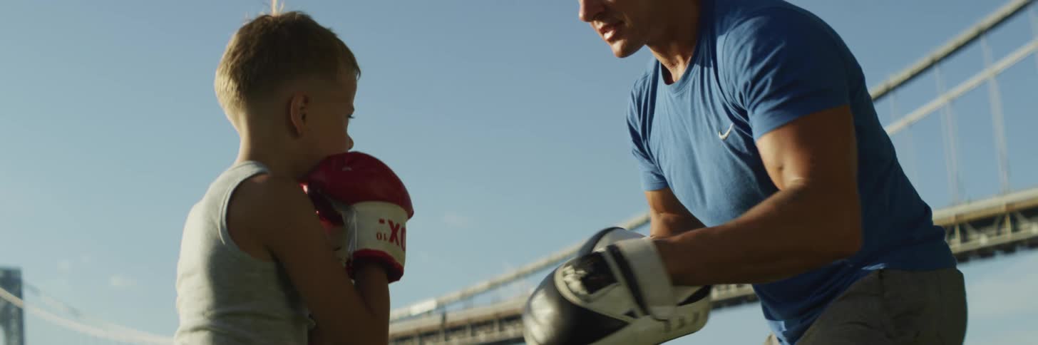 A boy wearing boxing gloves punches punch mitts being worn by a man as they stand outdoors near a large bridge on a clear day
