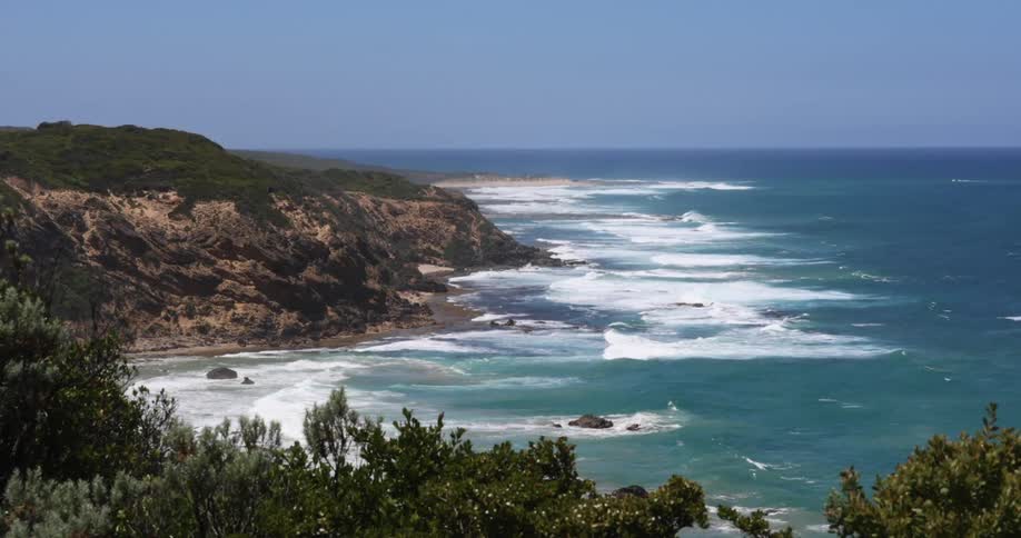 Ocean near horizon under misty sky is a darker blue than area near cliff as waves crash