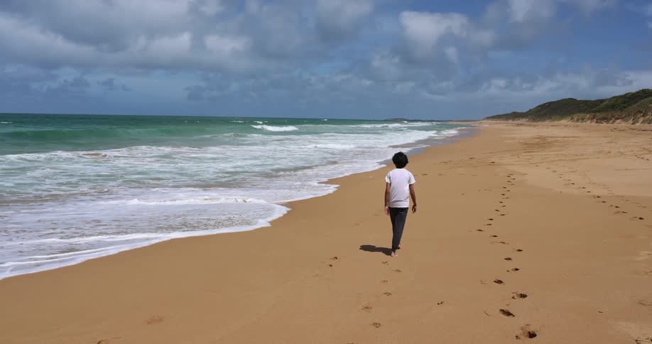 A boy walks barefoot on the beach leaving foot prints near the water