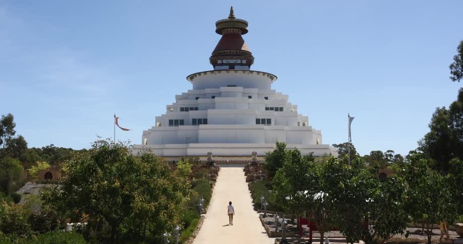 A person walks up a long slope lined with trees towards a large elaborately designed building looming over the surroundings