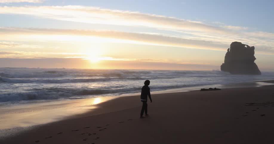 A person walking on the sand beach at sunset next to a rock promontory in the ocean