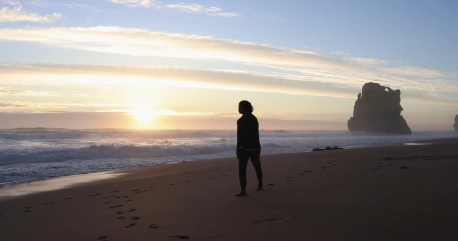 A teenager leaves footsteps in the sand while walking on the beach at sunset