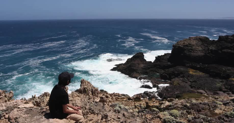 A young boy sits on a cliff on the shore and watches the waves rolling in