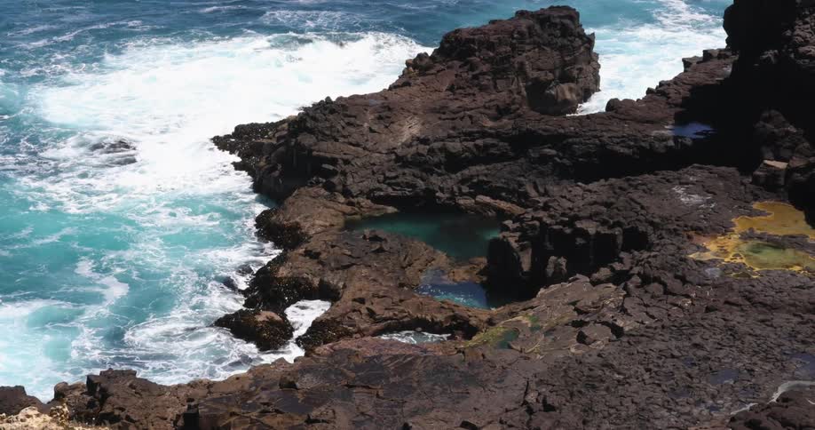 Wave crash against dark brown rocky shore dotted with pools tinted with algae