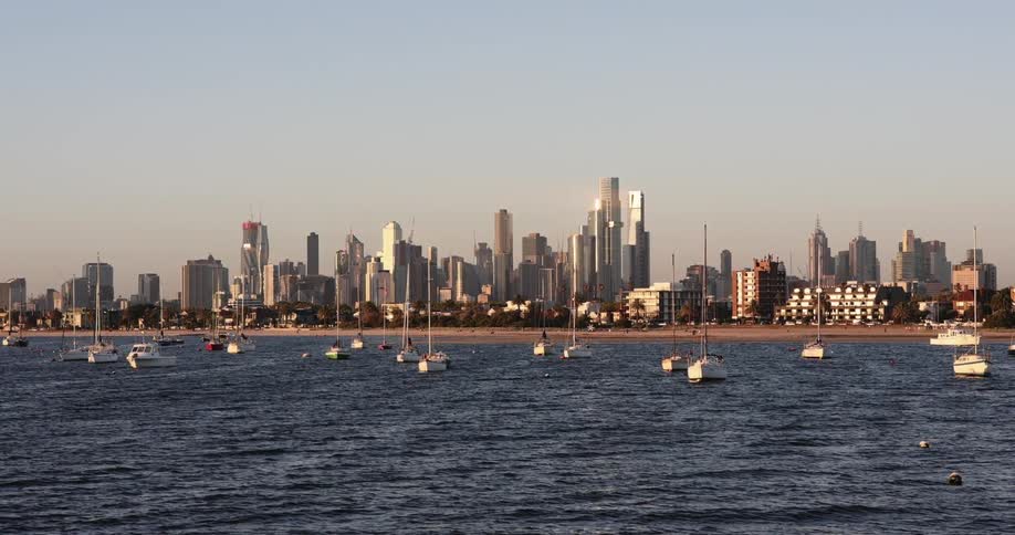 Sailboats with their sails down float in rows in water near beach bordering city