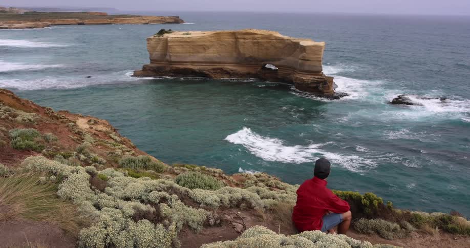A man sits along the edge of a promontory and watches waves move in the ocean below