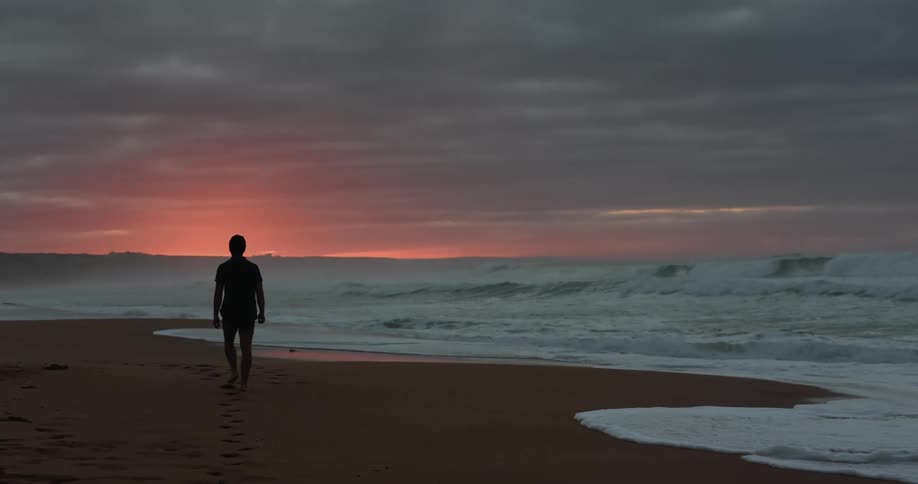 Man walks along beach toward red tinted sky leaving footprints