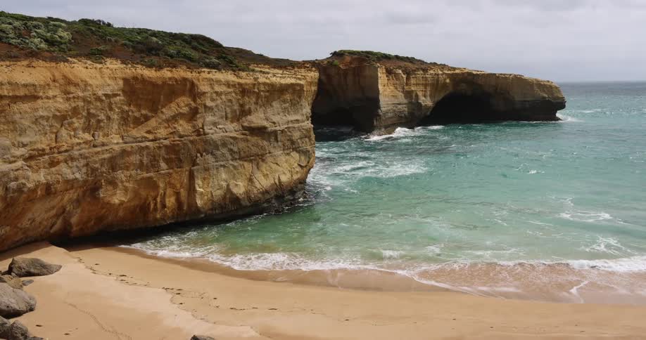 A beach with sand and waves next to a cliff with the sky and clouds in the background