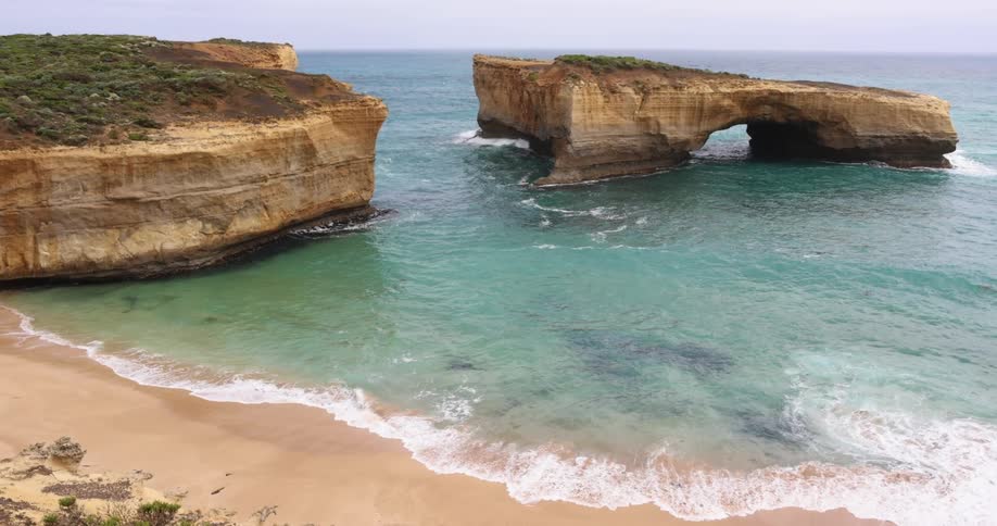 A beach with sand and waves next to a cliff with the sky and clouds in the background