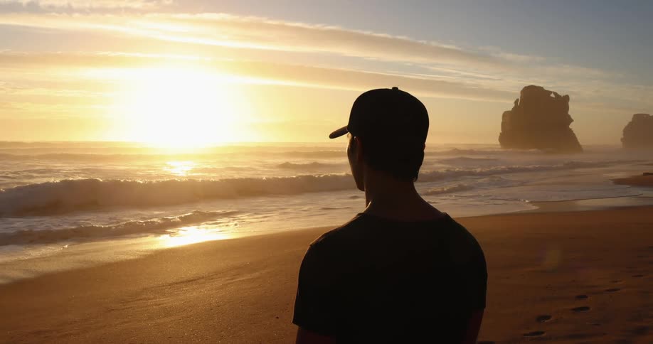 Man wearing a baseball cap looking at the sun setting over the ocean in a beach