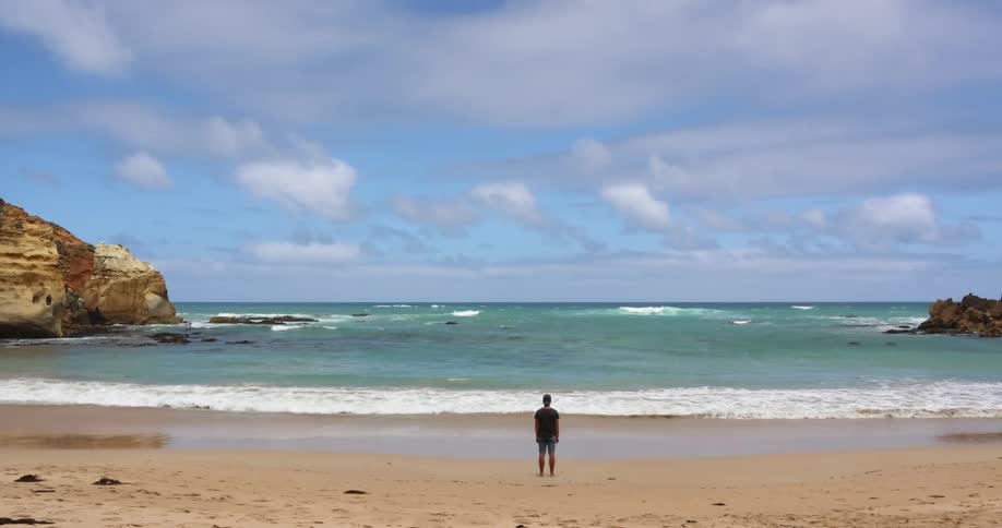 A man stands motionlessly on a beach facing the incoming tide on a sunny day