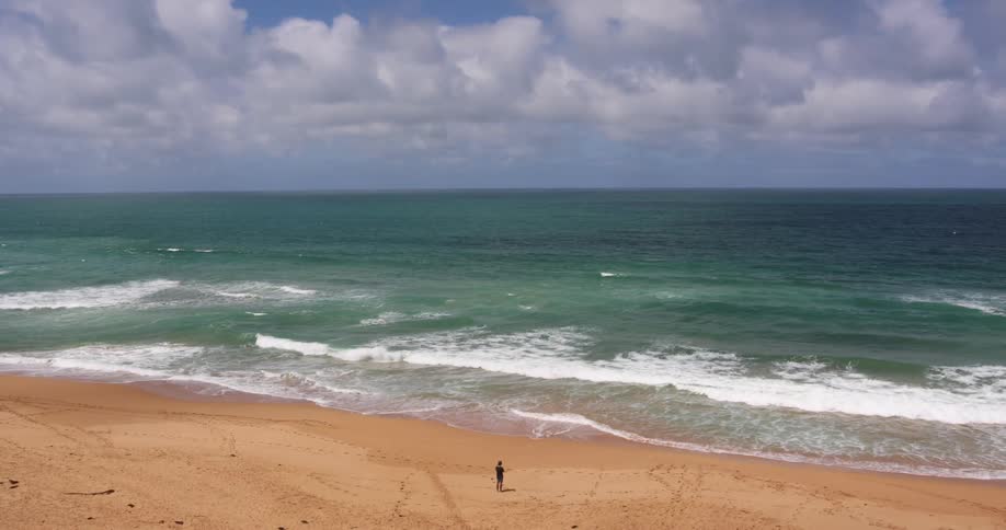 A person standing on a sandy beach with waves with the sky and clouds in the background