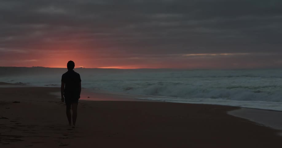 A person walking on a sandy beach with waves with the sky and clouds in the background